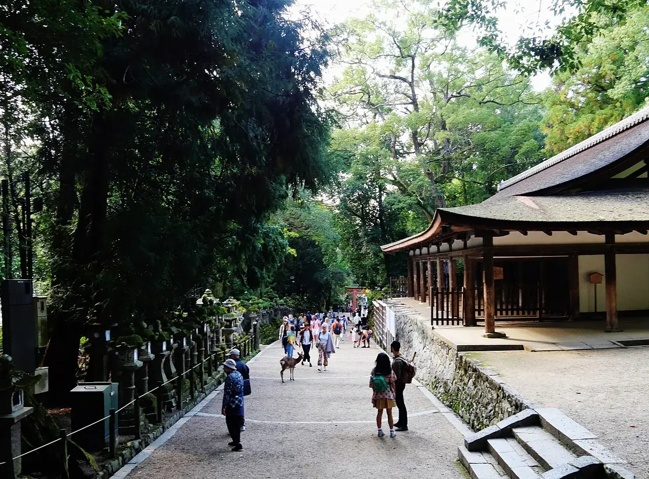 Stone lanterns lining the approach to Kasuga Taisha, with visitors and a deer on the path and a hall to the right
